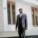 Stylish groom in blue suit posing outdoors in front of a modern building in India.